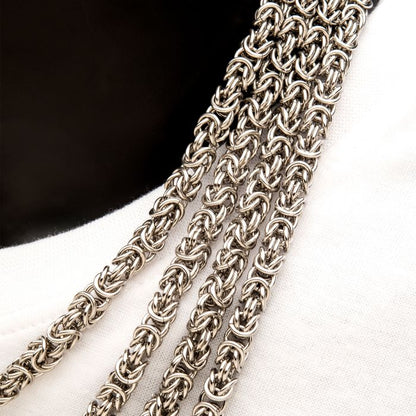 Close-up of four intricate silver chain necklaces laid over a white fabric surface with a black background. The chains feature the complex interwoven design of the INOX 6mm Steel King Byzantine Chain, showcasing their detailed craftsmanship. For those seeking durability, a stainless steel option is also available as the 22" INOX King Byzantine Chain.