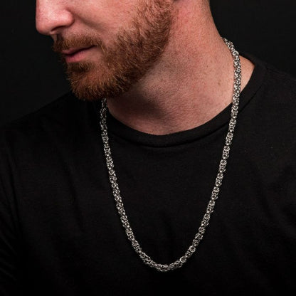 A man with a closely trimmed beard is wearing a black shirt and an INOX 6mm Steel King Byzantine Chain necklace. The background is dark, and the image focuses on his INOX chain necklace and part of his face, which is turned to the side.