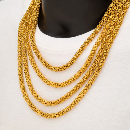 A mannequin showcasing a collection of layered gold chains over a white shirt features an eye-catching 6mm 18K Gold Plated King Byzantine Chain from INOX. The intricate link designs of the 20" chain create a bold and luxurious look, with the contrast between the vibrant gold and the crisp white shirt enhancing the jewelry's radiant shine.