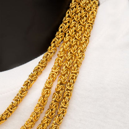 Close-up of a person wearing multiple strands of intricately woven 18K gold plated chains. The chains are draped over a white fabric, contrasting against the wearer's dark background. The golden links are detailed and shiny, highlighting the craftsmanship, reminiscent of the 6mm 18K Gold Plated King Byzantine Chain by INOX.