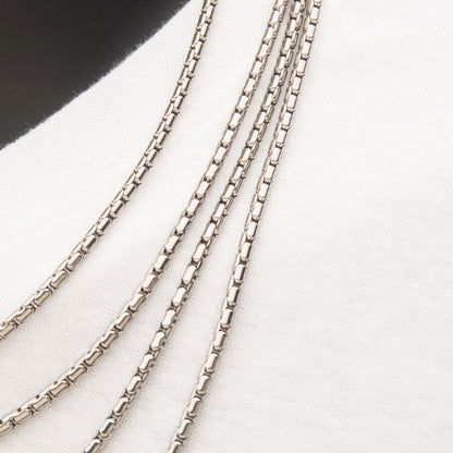 Close-up shot of four 3mm Steel Boston Link Chains | 24" by INOX, neatly laid out in a parallel arrangement on a white and black fabric background. These chains feature a detailed, geometric link pattern, showcasing their fine craftsmanship and shiny surface.