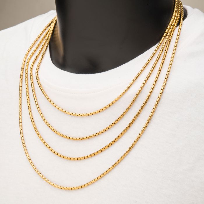 A person in a white shirt is wearing multiple layered 3mm 18K Gold Plated Boston Link Chain Necklaces by INOX around their neck. These finely crafted necklaces feature a delicate and consistent pattern. The photograph focuses on the neckline area, highlighting the elegance of the INOX gold jewelry.