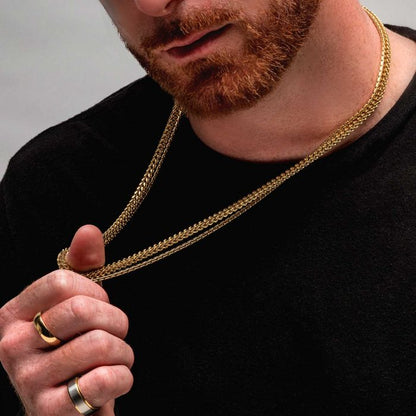 A bearded man wearing a black shirt and multiple rings holds the 3mm 18K Gold Plated Boston Link Chain by INOX around his neck with both hands. The focus is on the men's jewelry and the lower half of his face.