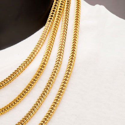 A close-up of three chains, including the 6mm 18K Gold Plated Foxtail Chain from INOX, draped over white fabric. The intricate, woven design of the chains showcases their shiny and polished appearance. The neutral background emphasizes the richness and detail of the foxtail chain and other exquisite designs.