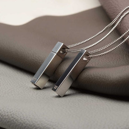 A close-up of two sleek, rectangular metallic pendants with smooth, reflective surfaces, suspended from delicate chains. Crafted from 925 sterling silver and engravable for personalization, The Keepsake Urn Engravable Pendant with Chain by INOX features a modern, minimalist design that stands out against a background of folded, textured fabrics in neutral tones.