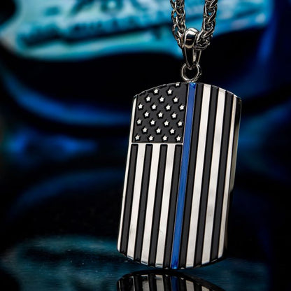 The INOX American Flag Police Officer Military Style Dog Tag Enamel Pendant with Chain is a silver necklace featuring the American flag design in black and white, accented by a single blue stripe running vertically on the right side to symbolize the thin blue line. This striking pendant stands out against a blurred background in shades of blue and black.