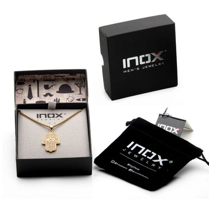 An 18K Gold Plated CZ Hamsa Pendant with a 22" Gold Plated Box Chain from INOX is displayed in an open black box featuring the "INOX" logo. The backdrop showcases various men's accessories. Nearby, there's a closed box and a small black drawstring pouch, both also branded "INOX".