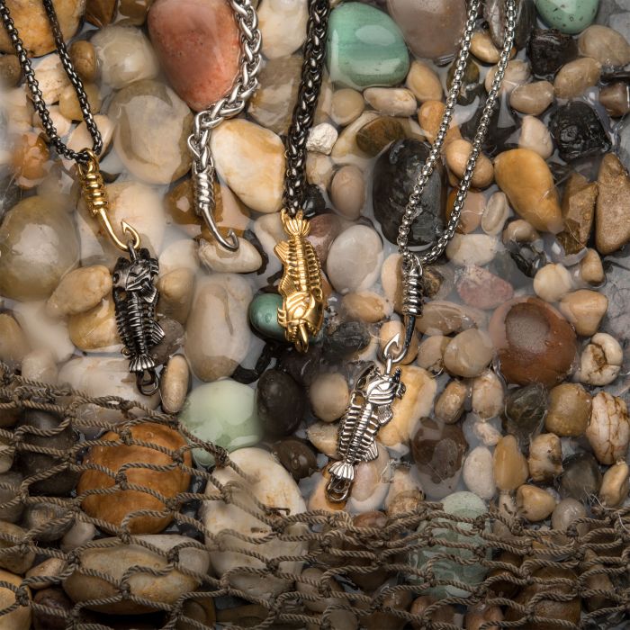 Four Black Plated Fishbone Pendants on Polished Gold-Plated Hooks with Black Plated Box Chains from INOX lie on gravel and pebbles, partially submerged in water. A fishing net is draped across the bottom of the image.