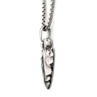 The Brushed Steel Chiseled Arrowhead Pendant with Box Chain by INOX features a silver men's pendant with a brushed steel finish, resembling an elongated, irregularly shaped, rough-textured arrowhead. The medium thickness chain sports a box link design. The handcrafted appearance of the arrowhead pendant highlights its unique, rugged allure.